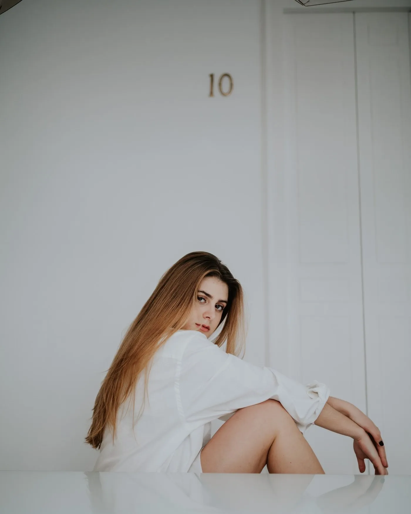 mujer sentada en una superficie blanca, camisa blanca oversized, mirando por encima del hombro con una expresión serena y directa. Luz suave y difusa, fondo minimalista. Fotografía de boudoir de Víctor Ruiz, Granada.