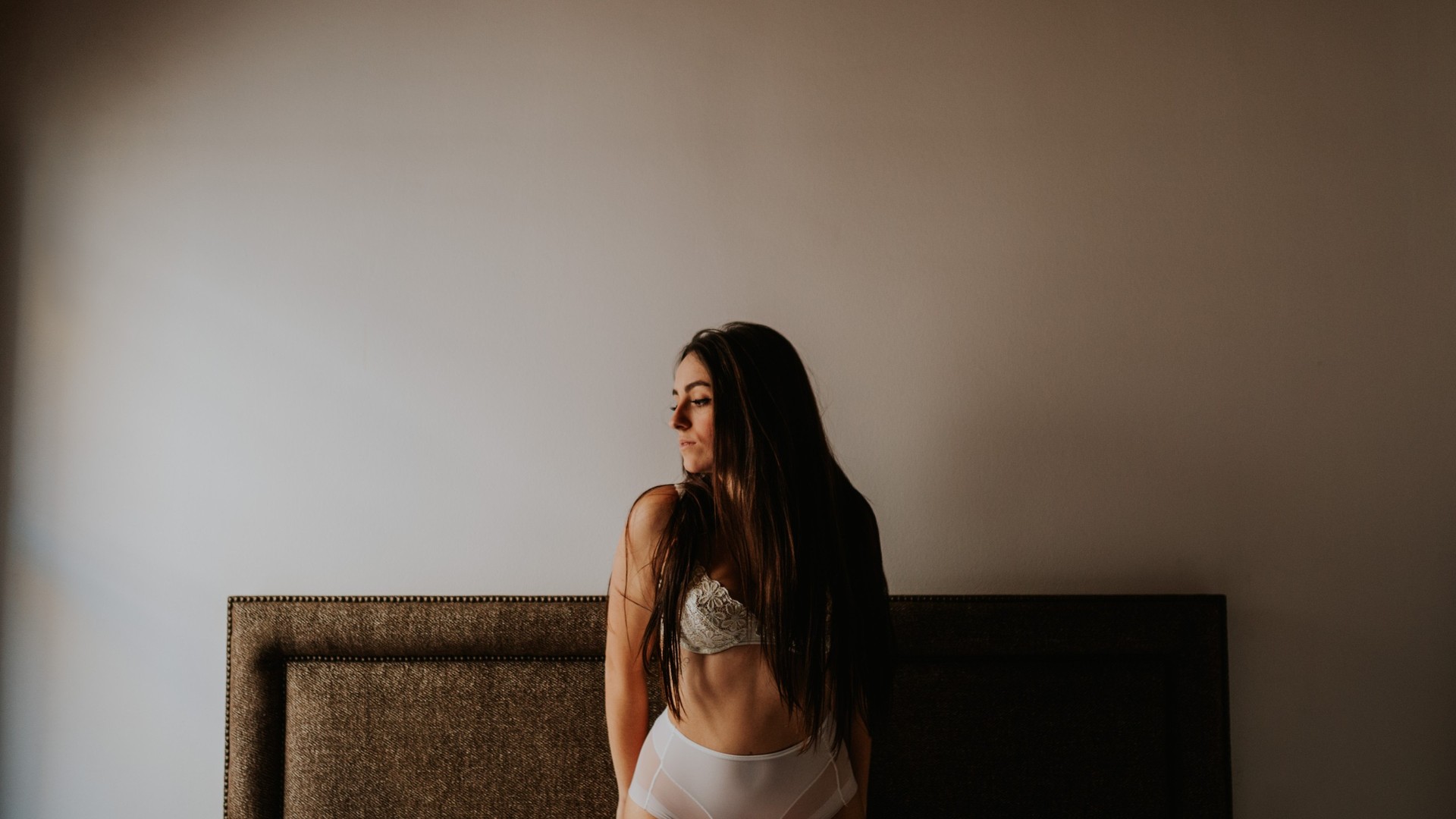 Mujer de cabello largo oscuro en lencería blanca de encaje, de pie frente a una cama con cabecero tapizado, iluminada por luz lateral natural suave. Fotografía de boudoir de Víctor Ruiz, Granada.