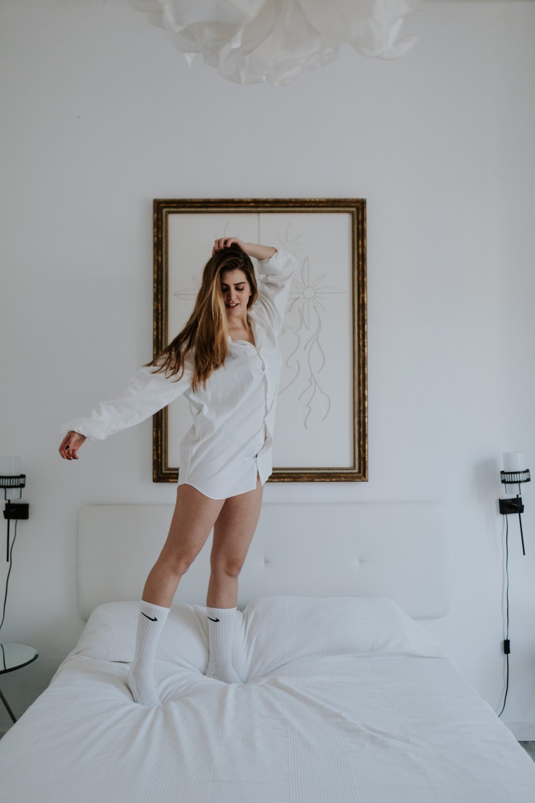 Mujer de pie sobre una cama blanca vistiendo una camisa blanca oversized, en un dormitorio minimalista bañado por luz natural. Fotografía de boudoir de Víctor Ruiz, Granada.
