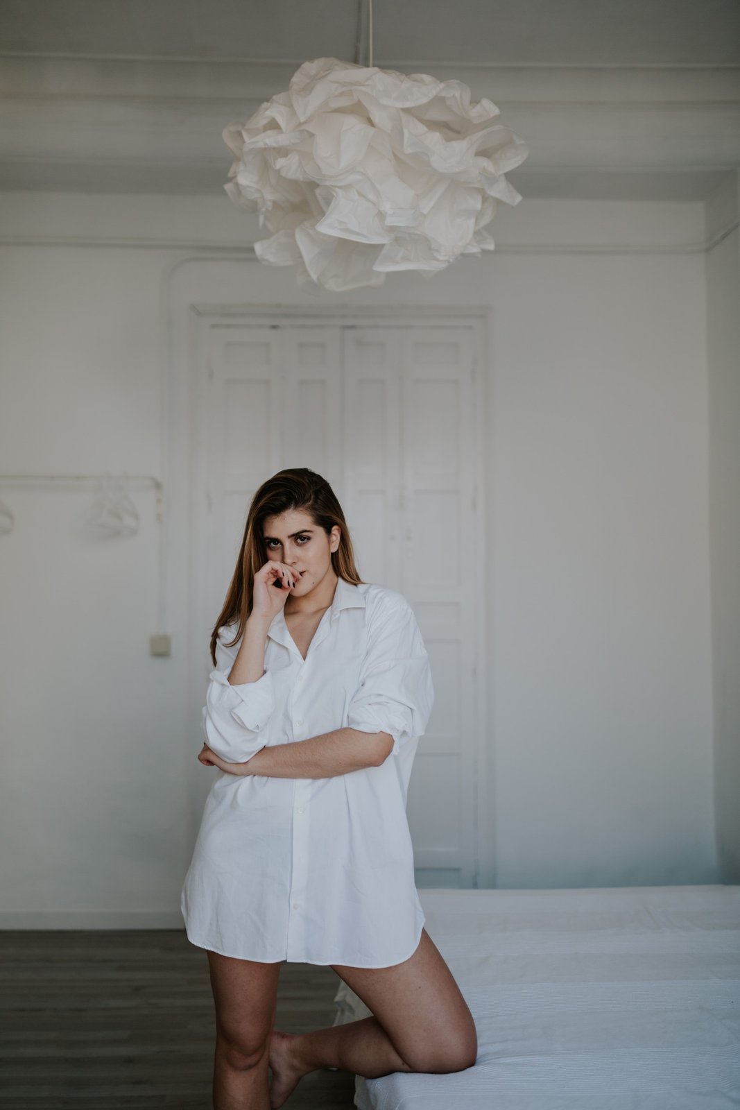 Mujer con camisa blanca holgada apoyada en el borde de una cama con una mano cerca de la boca, bajo una lámpara colgante escultórica blanca. Fotografía de boudoir de Víctor Ruiz, Granada.