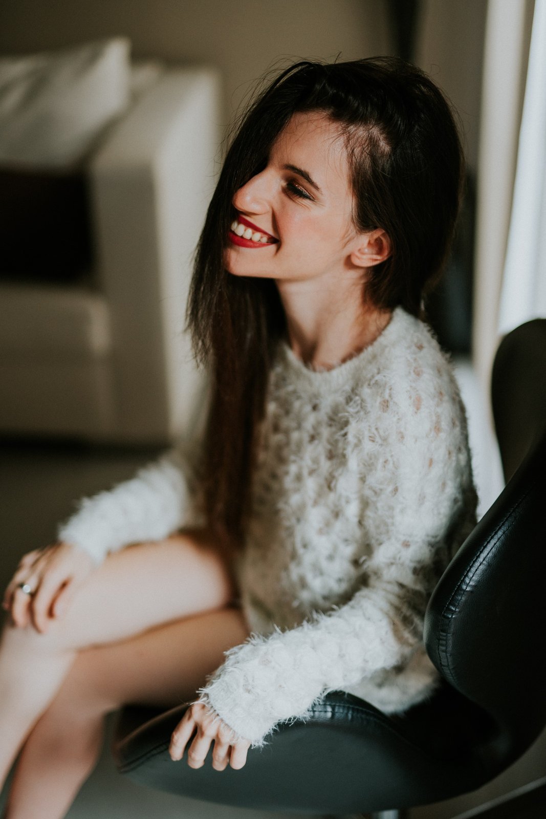 Mujer sentada en una silla con un suéter de punto crema, piernas cruzadas, sonriendo con el pelo oscuro suelto, iluminada por luz natural suave. Fotografía de boudoir de Víctor Ruiz, Granada.