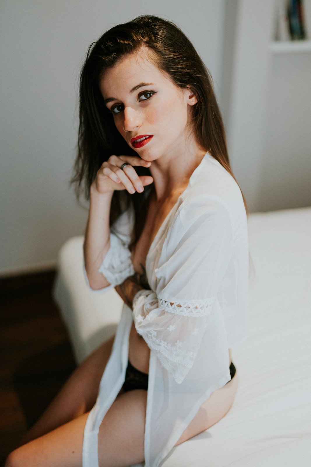 Mujer sentada al borde de una cama con sábanas blancas vistiendo una bata blanca translúcida con encaje en las mangas, con una mano en el mentón y mirada intensa a cámara. Fotografía de boudoir de Víctor Ruiz, Granada.