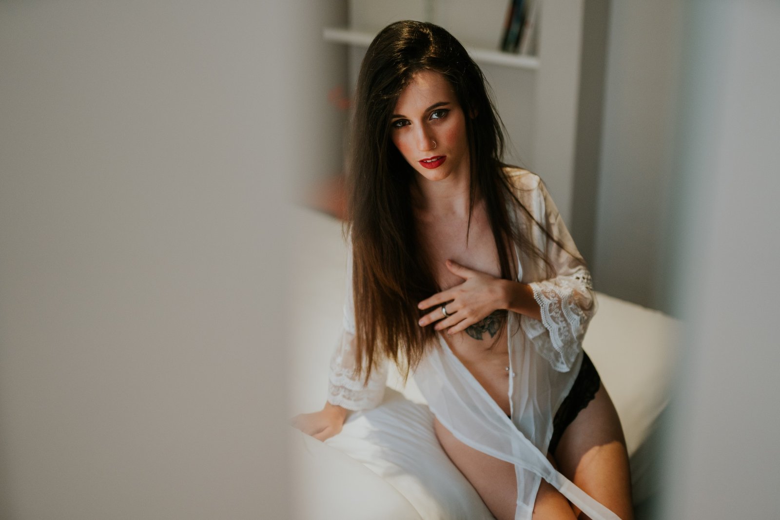 Mujer en una cama con sábanas blancas vistiendo una bata blanca abierta, recostada sobre las manos con una mano en el pecho y mirada suave. Fotografía de boudoir de Víctor Ruiz, Granada.