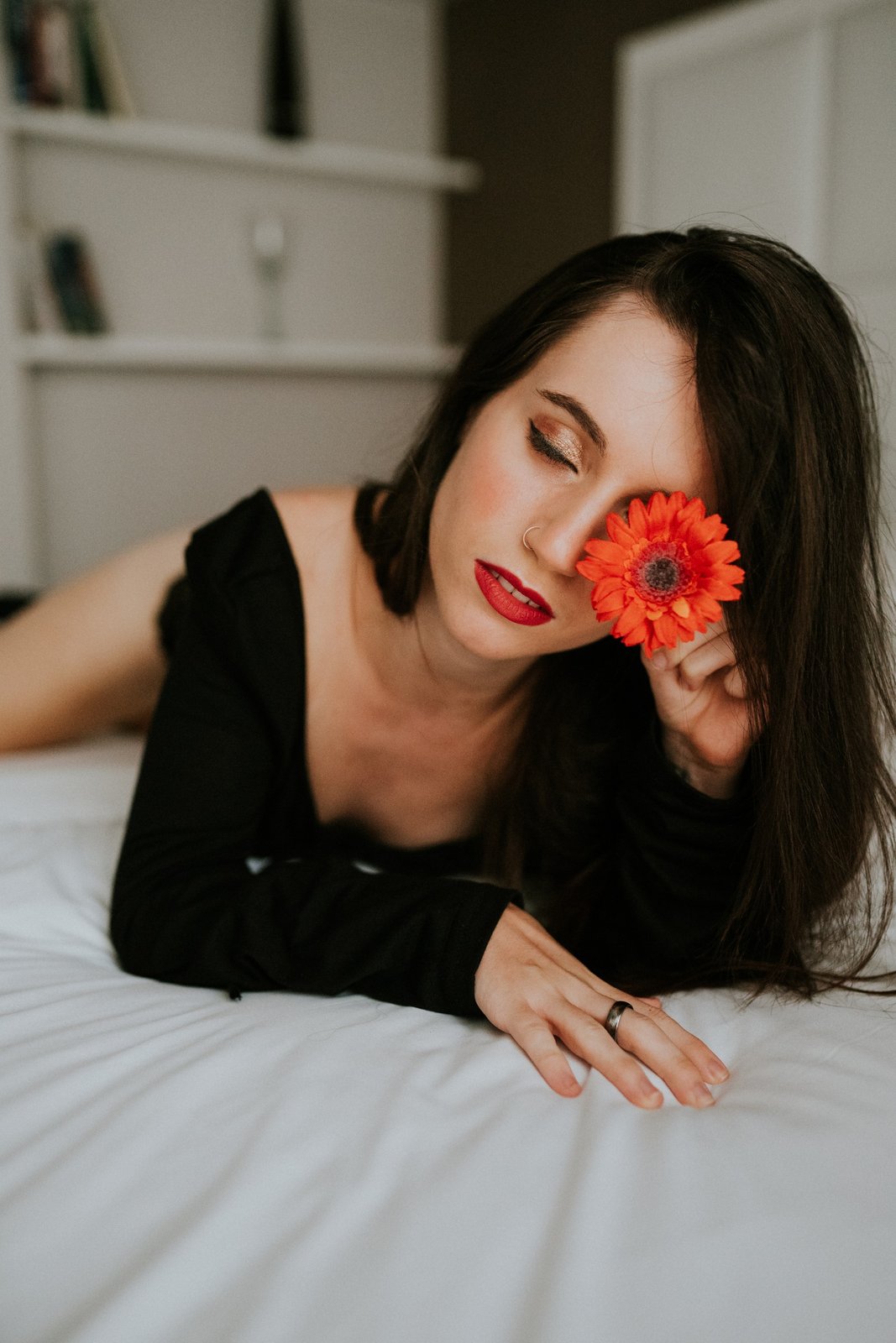 Primer plano de una mujer tumbada boca abajo con un top negro sin tirantes y labios rojos, sosteniendo una gerbera roja sobre uno de sus ojos. Fotografía de boudoir de Víctor Ruiz, Granada.