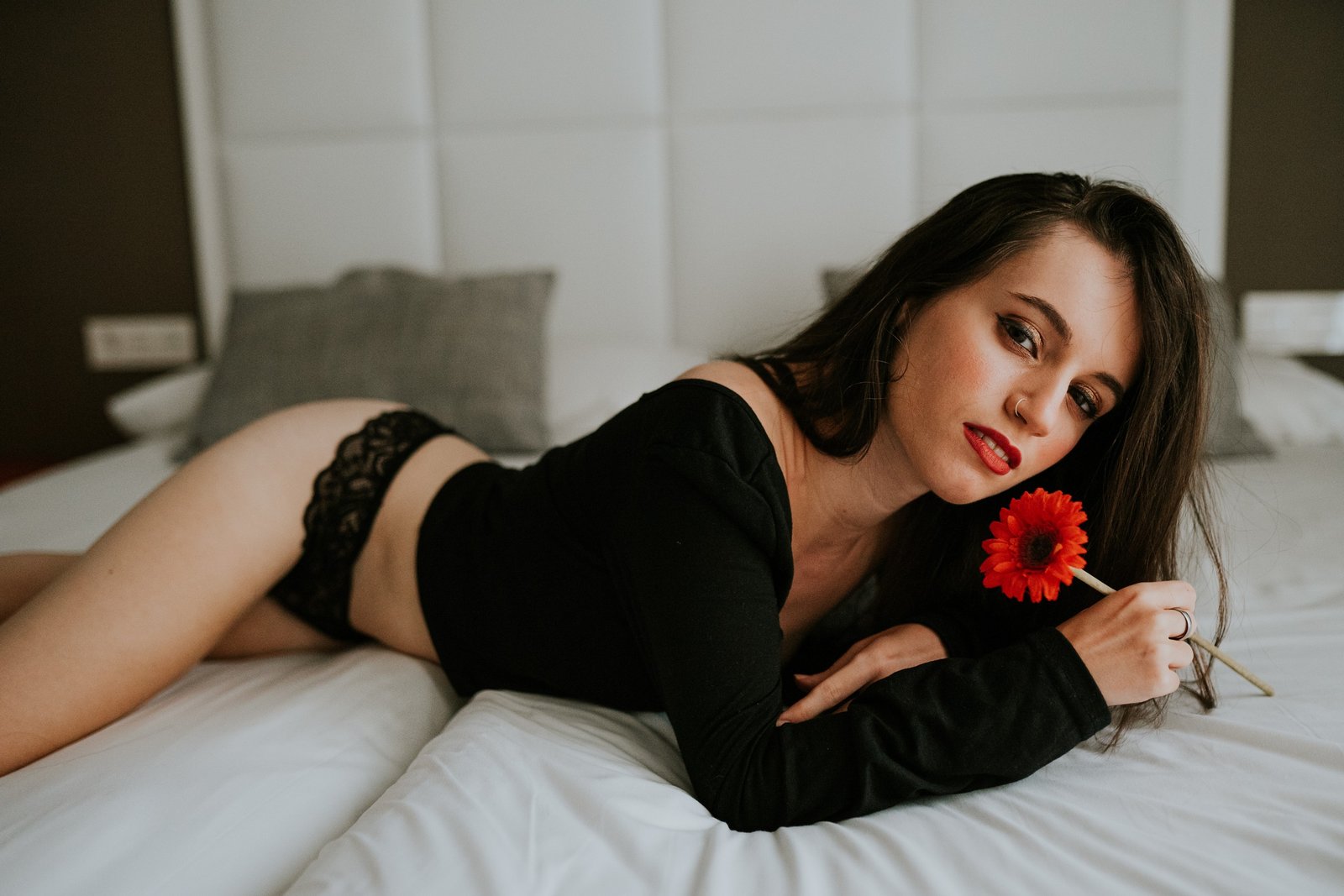 Mujer tumbada boca abajo en una cama de sábanas blancas con un top negro y ropa interior de encaje negro, sosteniendo una gerbera roja junto a su rostro y mirando a cámara. Fotografía de boudoir de Víctor Ruiz, Granada.