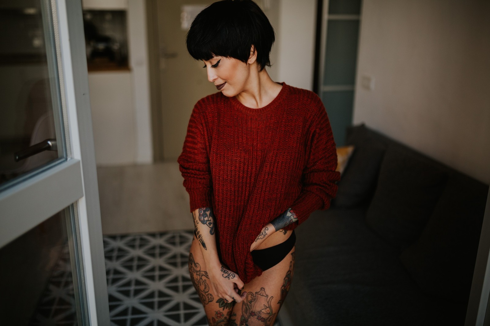 Mujer con tatuajes en un suéter rojo holgado y ropa interior negra, de pie en un umbral con luz natural, interior luminoso. Fotografía de boudoir de Víctor Ruiz, Granada.