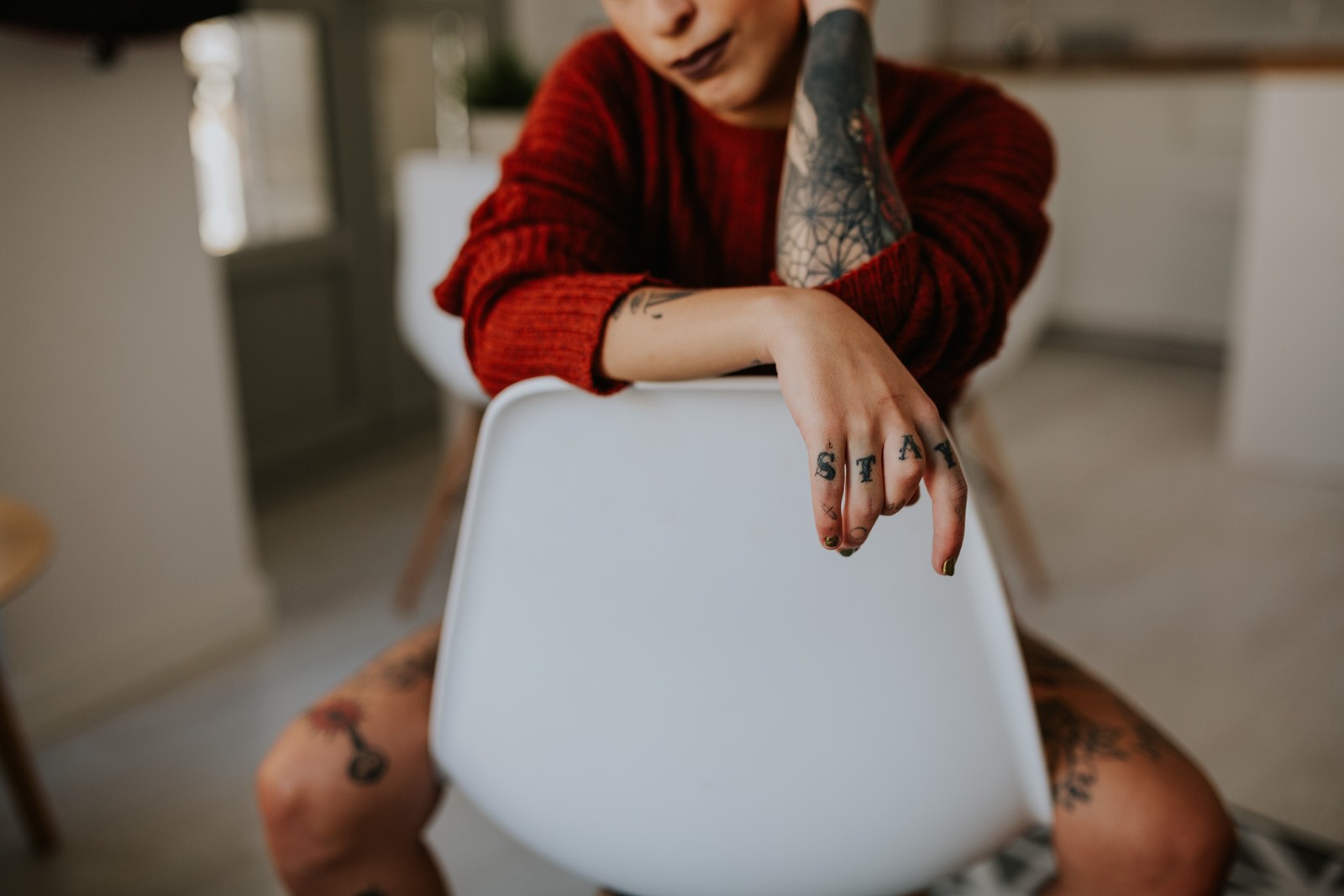 Persona con tatuajes extensos sentada en una silla blanca recostada hacia adelante con un suéter rojo de punto, luz natural suave. Fotografía de boudoir de Víctor Ruiz, Granada.