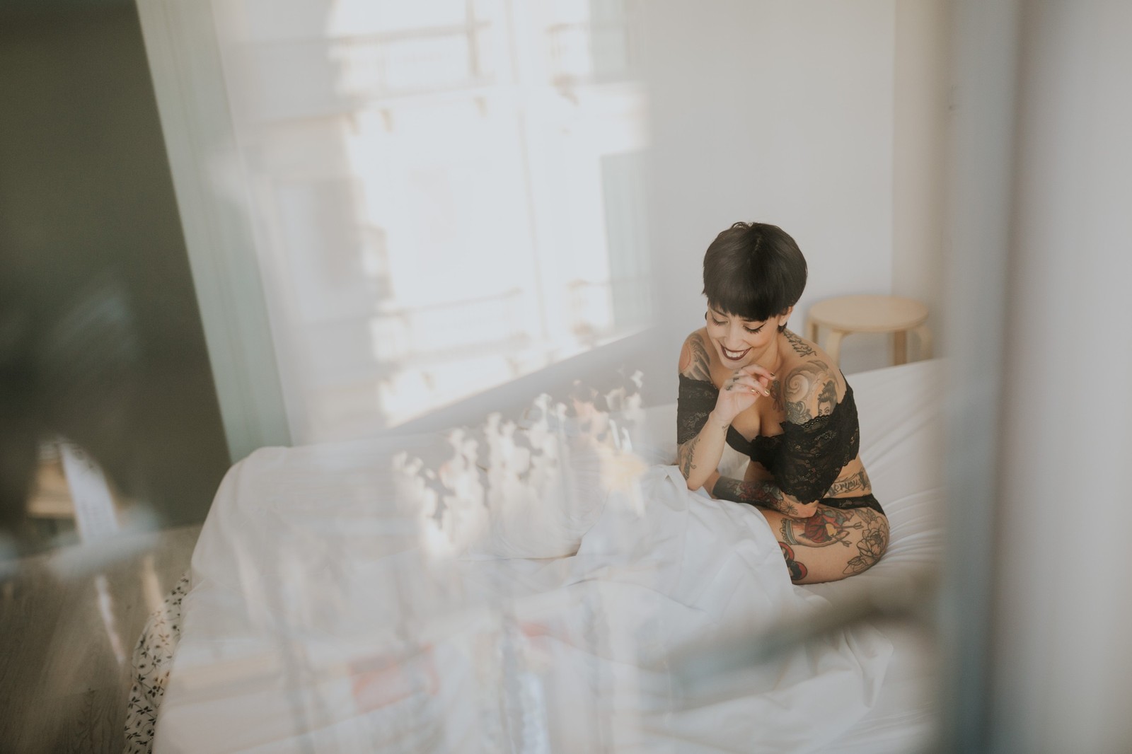 Mujer con cabello corto y tatuajes sentada en una cama sonriendo, en una habitación luminosa con luz natural suave. Fotografía de boudoir de Víctor Ruiz, Granada.
